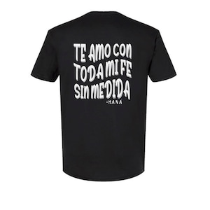 May include: Black t-shirt with white text on the back. The text reads "TE AMO CON TODA MI FE SIN MEDIDA -MANA" in a wavy, stylized font. The shirt has short sleeves and a crew neck.