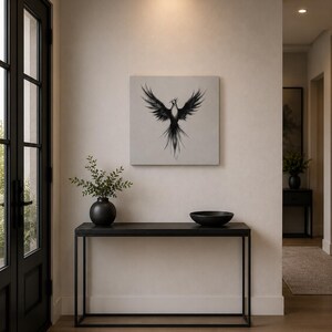 May include: A square wall art piece featuring a black ink illustration of a phoenix with wings spread wide, centered on a light gray background. The artwork is displayed above a black metal console table with a black vase and a black bowl.