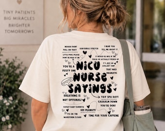 May include: A cream-colored t-shirt featuring black text on the back that reads NICU NURSE SAYINGS, surrounded by various phrases like NOOOO DONT PULL ON THAT, LITTLE POKE PLEASE, YAY SPA DAY, and HIII PEANUT, accented with small heart illustrations.
