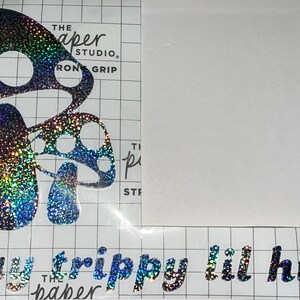 Könnte beinhalten: Ein schwarzer und silberner, schimmernder Vinyl-Aufkleber mit zwei Pilzen und dem Text "Stay trippy lil hippie" in einer kursivschrift.