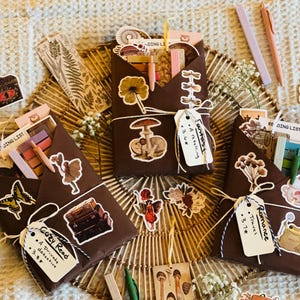 May include: Four brown paper parcels wrapped with twine and decorated with various stickers, including mushrooms, butterflies, and a typewriter. Each parcel has a small tag with handwritten text, including "Cozy Read", "Antares", "Romance", and "A Retelling".