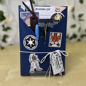 May include: A navy blue book sleeve with a white string closure, decorated with stickers of a tiger, a robot, and a Star Wars symbol. The sleeve contains a reading list, a pen, and a bookmark.