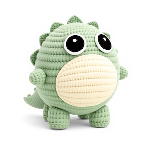 May include: A round, sage green crocheted dinosaur plush toy with a cream-colored belly, large black and white circular eyes, and small spikes running along its back. The creature has four short legs, two small arms, and a short tail.