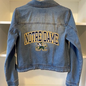 Puede incluir: Una chaqueta vaquera azul con un logotipo de Notre Dame en oro y verde en la parte posterior.