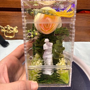以下が含まれることがあります： 透明なアクリルケースの中に、ミロのヴィーナスの白いミニチュア像、大きな淡いピンクの造花、緑の苔、そして様々な小さな造花が装飾品として配置されています。