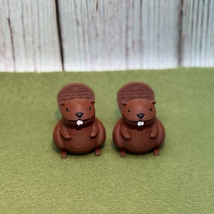 May include: Two brown beaver figurines with textured fur and large, flat tails. Each beaver has a small white tooth and tiny feet. The figurines are set against a green background with a wooden backdrop.