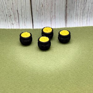 May include: Four small black pots filled with yellow material, possibly gold, are arranged on a green surface. The pots have a rounded shape and a small rim. The background is a light green surface and a wooden wall.