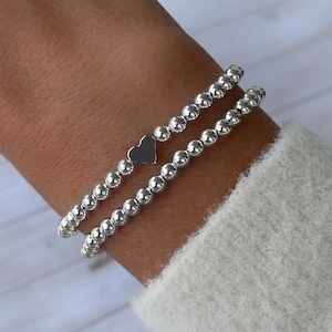May include: Two silver beaded bracelets stacked on a wrist. The top bracelet has a small silver heart charm.