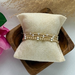 May include: Three gold and silver beaded bracelets are displayed on a beige linen pillow inside a small wooden bowl. The bracelets have varying sizes of gold and silver beads. Pink and white flowers are visible in the background.
