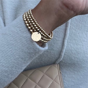 May include: A set of five gold beaded bracelets with a hammered gold disc charm. The bracelets are stacked on a person's wrist.