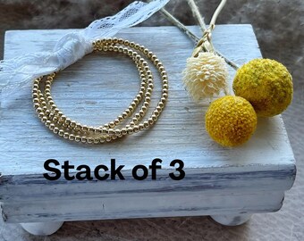 May include: A stack of three gold-toned beaded bracelets featuring small, uniform metallic spheres. The bracelets are tied together with a white lace ribbon and are displayed alongside two yellow and one cream-colored dried flower heads. Text reads: Stack of 3.