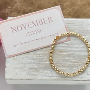 May include: A gold beaded bracelet with citrine stones. The bracelet is on a white wooden surface with a card that says "November Citrine" and lists the associated keywords: "creativity, success, optimism, energy".