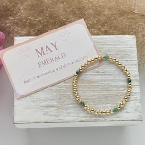 May include: A gold beaded bracelet with green emerald stones. The bracelet is on a white wooden surface with a card that says "MAY EMERALD balance ♥ optimism ♥ wisdom ♥ creativity".