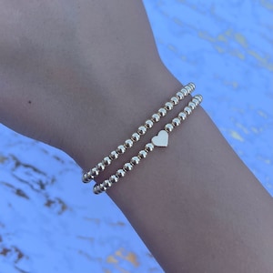 May include: Two gold beaded bracelets with a small gold heart charm. The bracelets are stacked on a person's wrist.