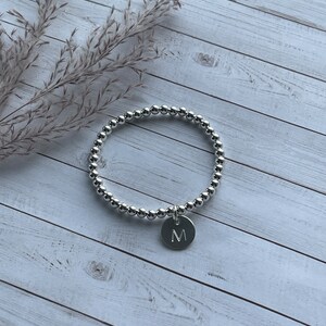 May include: A silver-toned beaded bracelet featuring a circular charm engraved with the letter M. The bracelet is constructed from a continuous strand of small, polished metallic spheres, creating a uniform and reflective circular design.