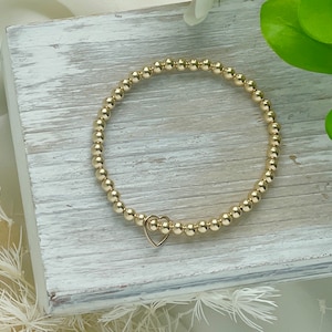 May include: A gold beaded bracelet with a small heart charm. The bracelet is on a white wooden surface.