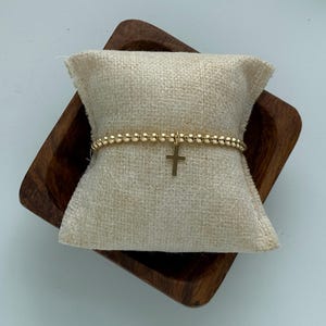 May include: A gold beaded bracelet with a small gold cross charm. The bracelet is displayed on a small wooden cushion.