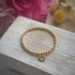 May include: A gold beaded bracelet with a small round charm. The charm has the letter "B" engraved on it. The bracelet is on a light-colored wooden surface, with pink flowers in the background.