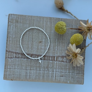 May include: A silver beaded bracelet with a small silver cross charm. The bracelet is on a wooden surface.