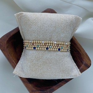 May include: Gold beaded bracelet with small, colorful accent beads in blue, green, and pink. The bracelet is displayed on a small, square, beige pillow inside a wooden bowl. The beads are small and round, creating a delicate and elegant look.