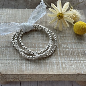 May include: Three silver bead bracelets stacked on a wooden surface. The bracelets are made of small, round beads and are a popular style of jewelry.