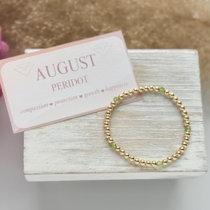 May include: A gold beaded bracelet with green peridot stones. The bracelet is on a white wooden surface with a pink card that says "August Peridot" and "compassion, protection, growth, happiness".
