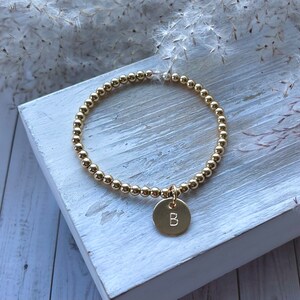 May include: A gold beaded bracelet with a gold charm that has the letter "B" engraved on it.