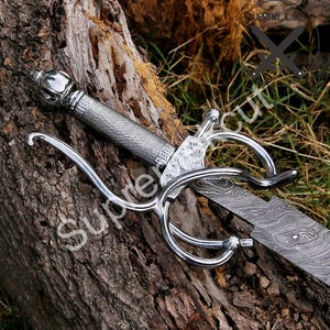 May include: A decorative sword with a silver handle and guard, and a patterned blade. The sword is lying on a piece of wood, with grass in the background. The handle has a textured grip.