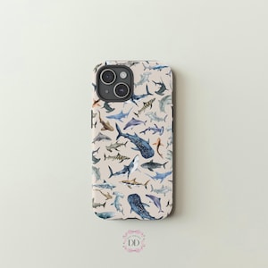 May include: A phone case featuring a pattern of various shark illustrations in shades of blue, gray, and brown against a beige background. The case has a camera cutout and a rounded shape.