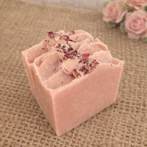 May include: A square, light pink bar of soap with a textured top, sprinkled with dried rose petals. The soap is set on a burlap surface, with a blurred background of pink roses.