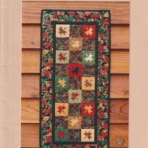 May include: A quilting pattern for a table runner featuring a design of tumbling leaves in shades of red, green, brown, and beige. The pattern is titled "Tumbling Leaves Runner, A Paper Foundation Pattern, MH Designs, MH 613".
