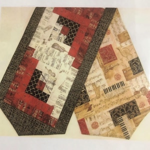 May include: A rectangular table runner featuring a patchwork design with musical motifs. The fabric displays sheet music, piano keys, violins, and harps in shades of red, black, cream, and tan. It has a dark brown textured border.