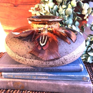 May include: A decorative, round vase with a speckled gray and brown finish, adorned with brown and red feathers. The vase has a gold-colored rim and sits atop a stack of blue books. The text "LONGFELLOW'S Evangeline" is visible on one book.
