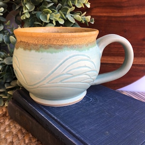 May include: A ceramic mug with a rounded body and a single handle. The mug features a light blue glaze on the lower portion with embossed wave-like patterns, transitioning to a speckled golden-brown glaze around the upper rim and interior.