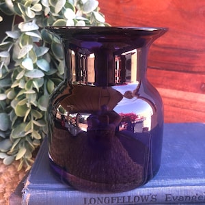 May include: A dark purple glass vase with a wide base and flared rim. The vase reflects the surrounding environment, including greenery and a wooden surface. The vase is sitting on a blue book.