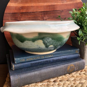 May include: A large, white ceramic bowl with a green and blue glaze. The bowl has a wavy rim and is sitting on a stack of blue books. The books have gold lettering on their spines, including "The Merchant of Venice" and "Longfellow's Evangeline".