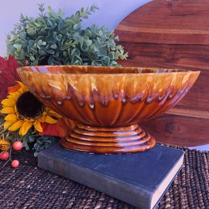 May include: A large, oval-shaped ceramic bowl with a pedestal base. The bowl has a glossy, brown glaze with lighter streaks. The base features concentric rings. The bowl is displayed on a dark blue book, with a wooden cutting board in the background.
