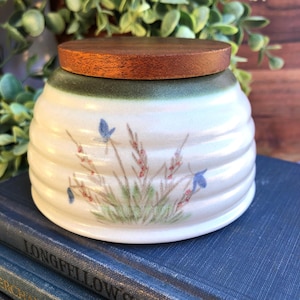 May include: A ceramic jar with a wooden lid. The jar is white with a ribbed design and a painted floral pattern in blue, red, and green. The lid is a dark brown color.