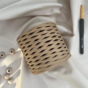 May include: A roll of tan-colored raffia ribbon with a woven design. A black and gold crochet hook is positioned to the right of the ribbon. Several small, light-colored wooden beads are scattered on the white fabric.