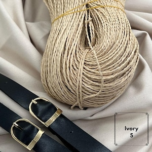May include: A large, coiled bundle of light beige twisted paper cord secured with a thin yellow rubber band, positioned next to two black leather-like straps featuring gold-toned metal buckles. The text Ivory 5 is printed in the lower right corner.