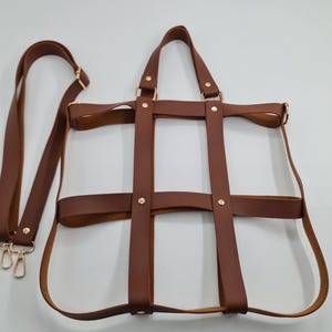 May include: A brown leather tote bag with a unique cage-like design. The bag features a long, adjustable shoulder strap and two shorter handles. Gold-colored hardware accents the bag's design. The bag is empty and sits on a white surface.