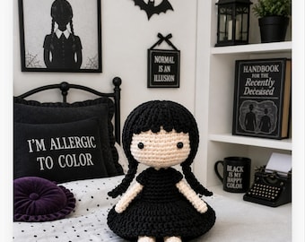 Può includere: Bambola all'uncinetto con capelli neri e un vestito nero. La bambola è seduta su un letto con un cuscino con la scritta "I'M ALLERGIC TO COLOR". Un'immagine incorniciata di "WEDNESDAY ADDAMS" è appesa al muro.