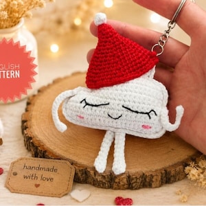 May include: A handmade crochet keychain featuring a white cloud-shaped design with a red Santa hat. The cloud has black eyes, pink cheeks, and white dangling arms and legs. The keychain is on a silver ring.