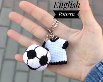 May include: A white and black crocheted soccer ball and a white crocheted t-shirt with black trim attached to a silver keychain. The keychain is held in a hand. The text "English Pattern" is above the keychain.