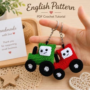 Può includere: Due portachiavi a forma di trattore fatti a mano all'uncinetto, uno verde e uno rosso, con ruote nere e dettagli bianchi. I portachiavi sono tenuti da una persona. Una carta dice "Handmade with love". Il testo "English Pattern" è anche visibile.