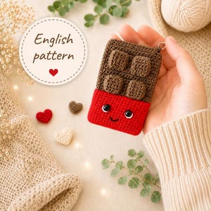 May include: A crocheted chocolate bar keyring with a smiling face and a red wrapper. The chocolate section is brown with square details. The image also includes crocheted hearts and the text "English pattern".