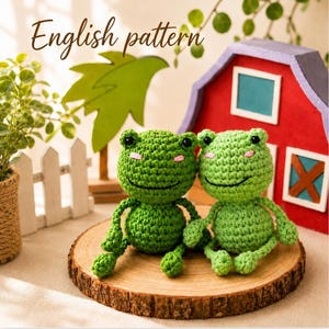 Op de afbeelding: Twee gehaakte kikkers met lichamen van groen garen, zwarte kraaloogjes en roze geborduurde wangen. De tekst "English pattern" is zichtbaar boven de kikkers, die op een ronde houten schijf voor een rood schuurgebouw zijn geplaatst.