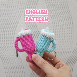 Op de afbeelding: Twee gehaakte drank sleutelhangers, één roze en één lichtblauw, met witte en grijze accenten. Elke sleutelhanger heeft een handvat en een zilveren ring. De tekst "ENGLISH PATTERN" staat erboven.