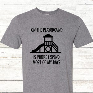 May include: A gray t-shirt with the text "ON THE PLAYGROUND IS WHERE I SPEND MOST OF MY DAYS" in black. A black graphic of a playground structure is centered below the text.