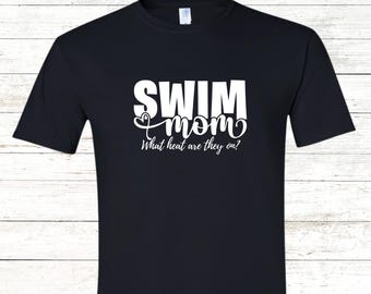 May include: Black t-shirt with the words "SWIM mom" in white block letters and script. Below the text is the phrase "What heat are they on?" in white script. The shirt is made of a soft material.